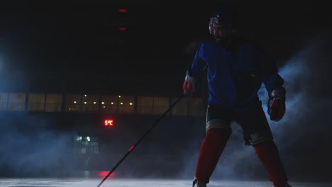 Man-hockey-player-with-a-puck-on-the-ice-in-hockey-form-leaves-with-a-stick-in-his-hands-out-of-the-darkness-and-looks-straight-into-the-camera